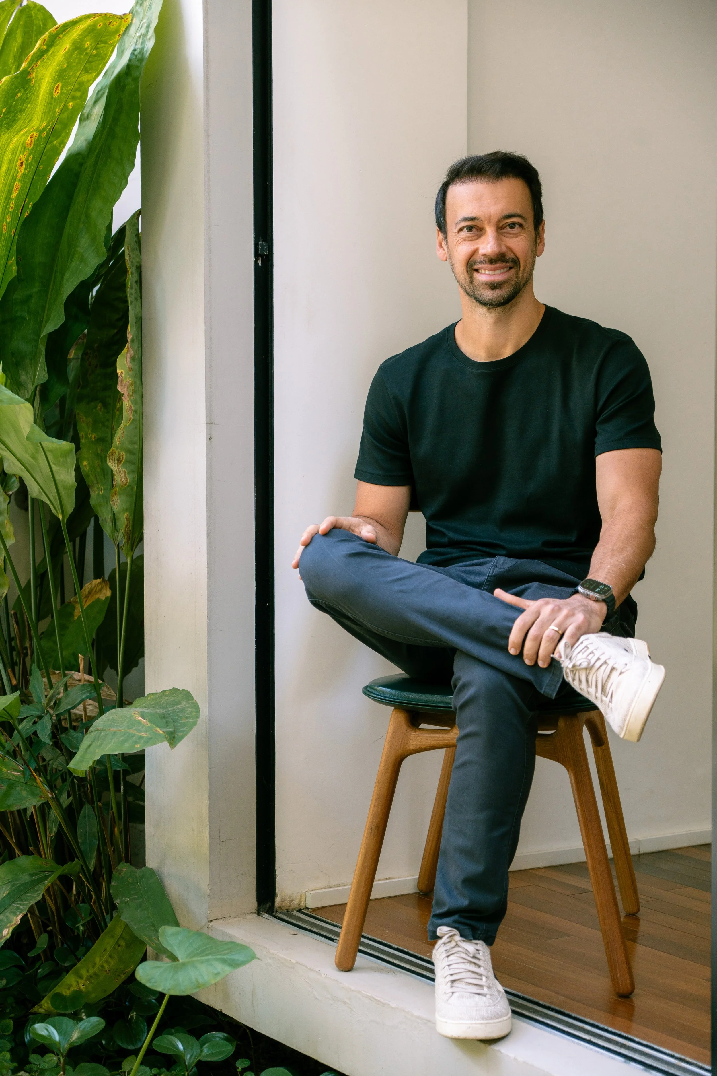 A smiling man with a beard, wearing a black T-shirt, blue pants and white sneakers, sitting on a wooden chair near a window, with green plants beside him.