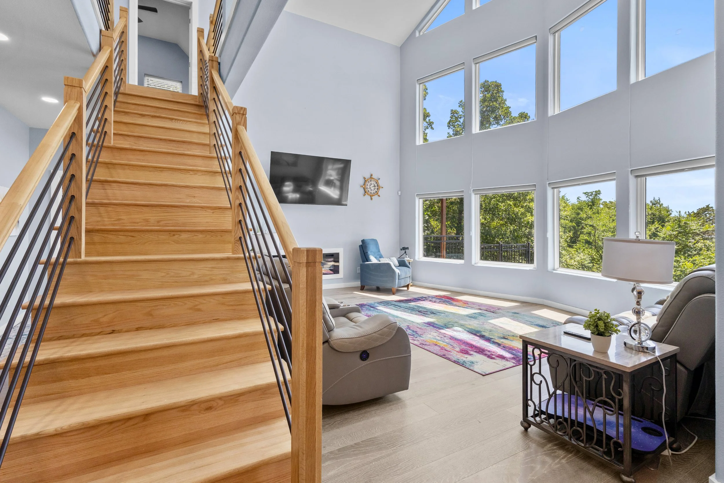 Bright living room with large windows, wooden staircase with black metal railing, two blue chairs, gray recliner, black side table, colorful rug, and TV mounted on white wall, with trees visible outside.
