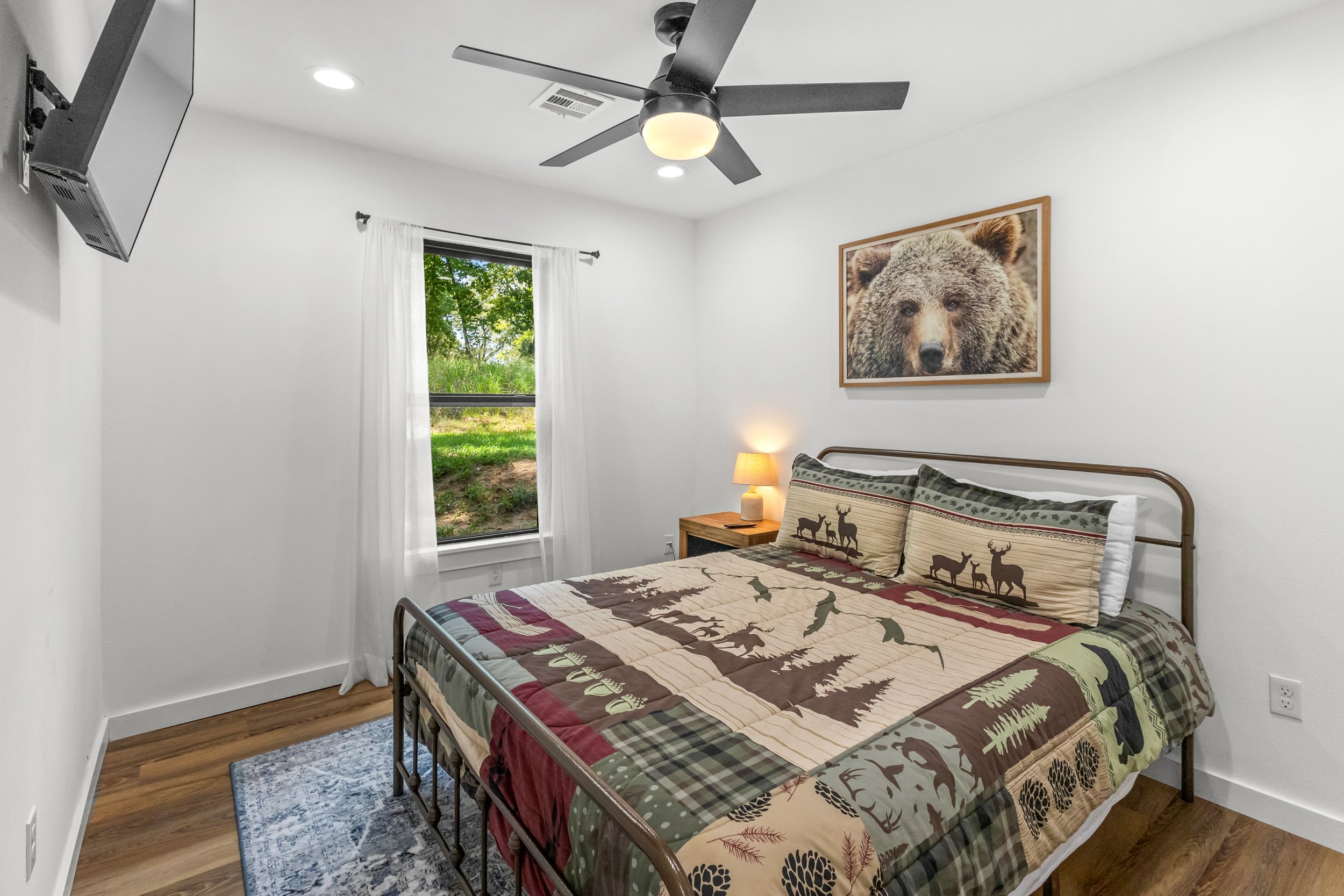 A bedroom with a bed, a nightstand with a lamp, a window with white curtains, and a picture of a bear on the wall.