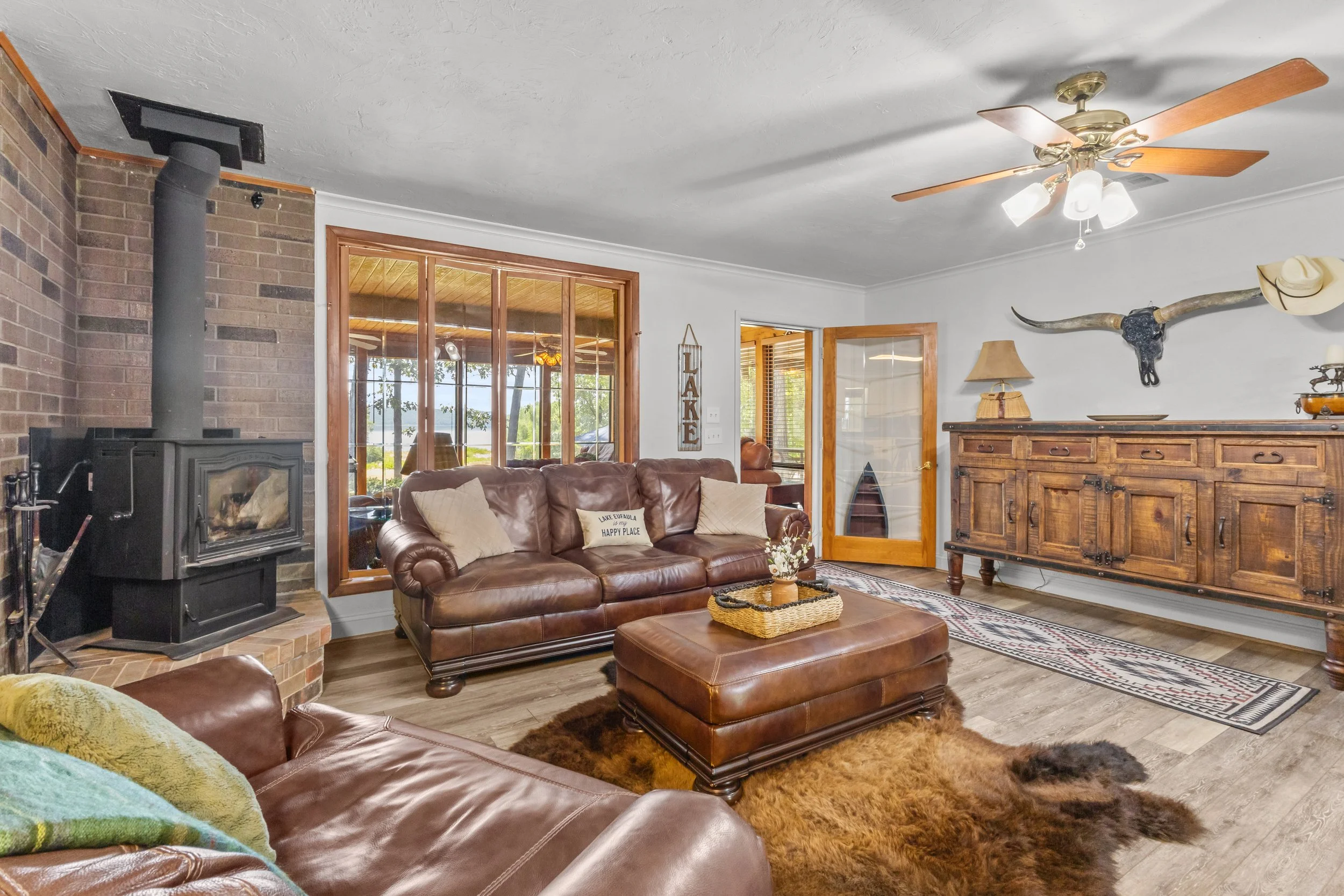 Living room with a leather sofa, a wood coffee table, a wood sideboard, a mounted longhorn skull with a cowboy hat, a ceiling fan, and a wood-burning stove near a large window showing a lake view.
