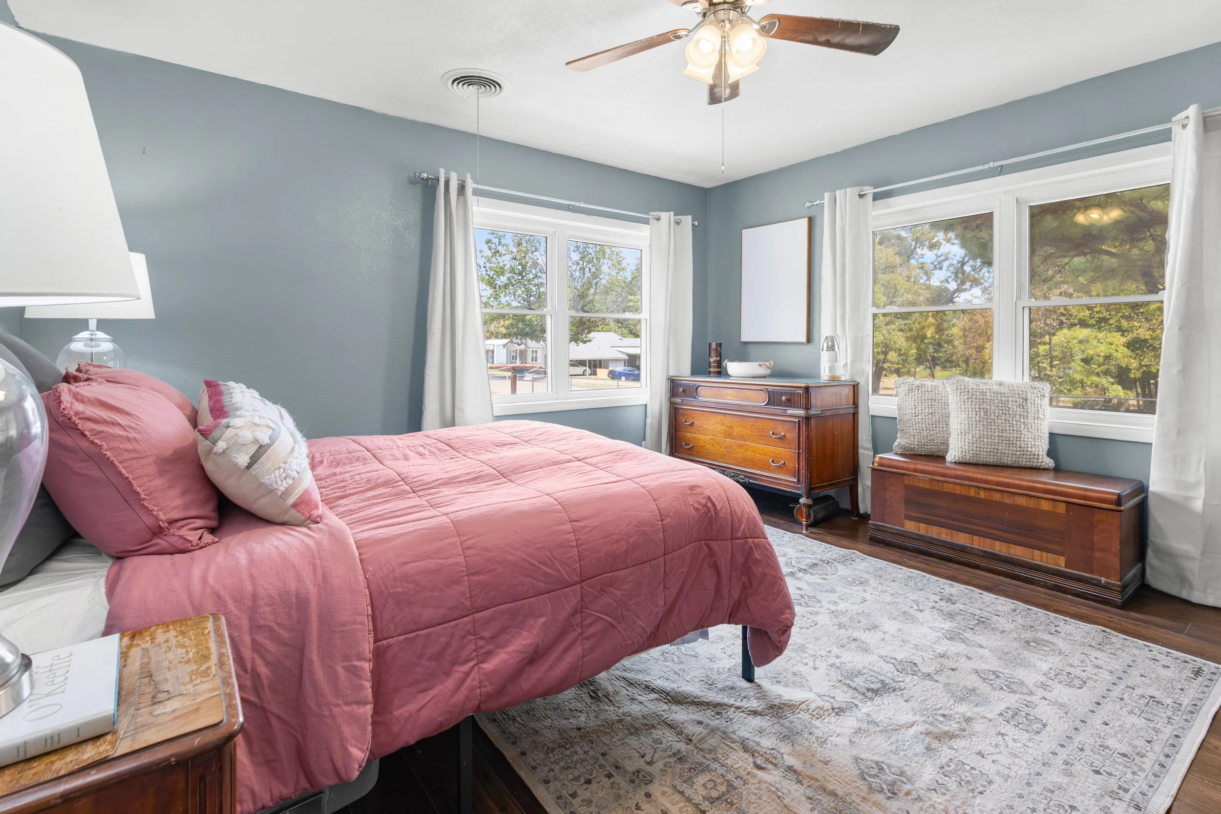 A cozy bedroom with a pink quilted bedspread, several pillows, wooden furniture, and large windows showing trees outside, illuminated by natural light and ceiling fan lights.