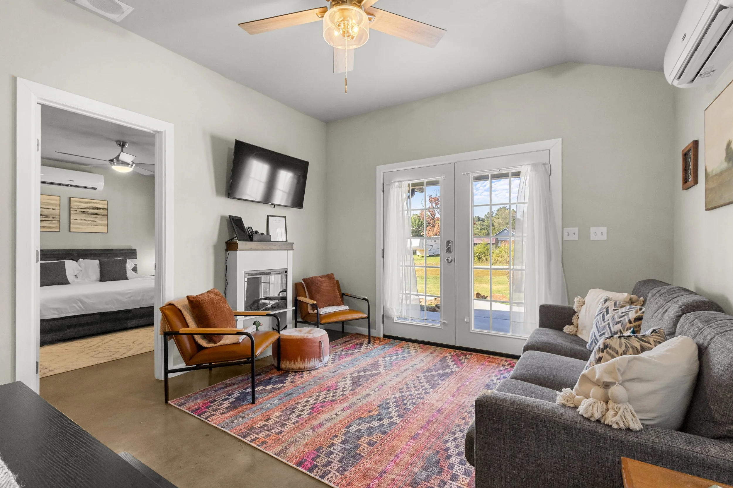 Living room with a gray sofa, two orange chairs, a fireplace, a large colorful rug, and a glass door leading outside. A TV is mounted on the wall, and a bedroom with a bed and ceiling fan is visible through a doorway.