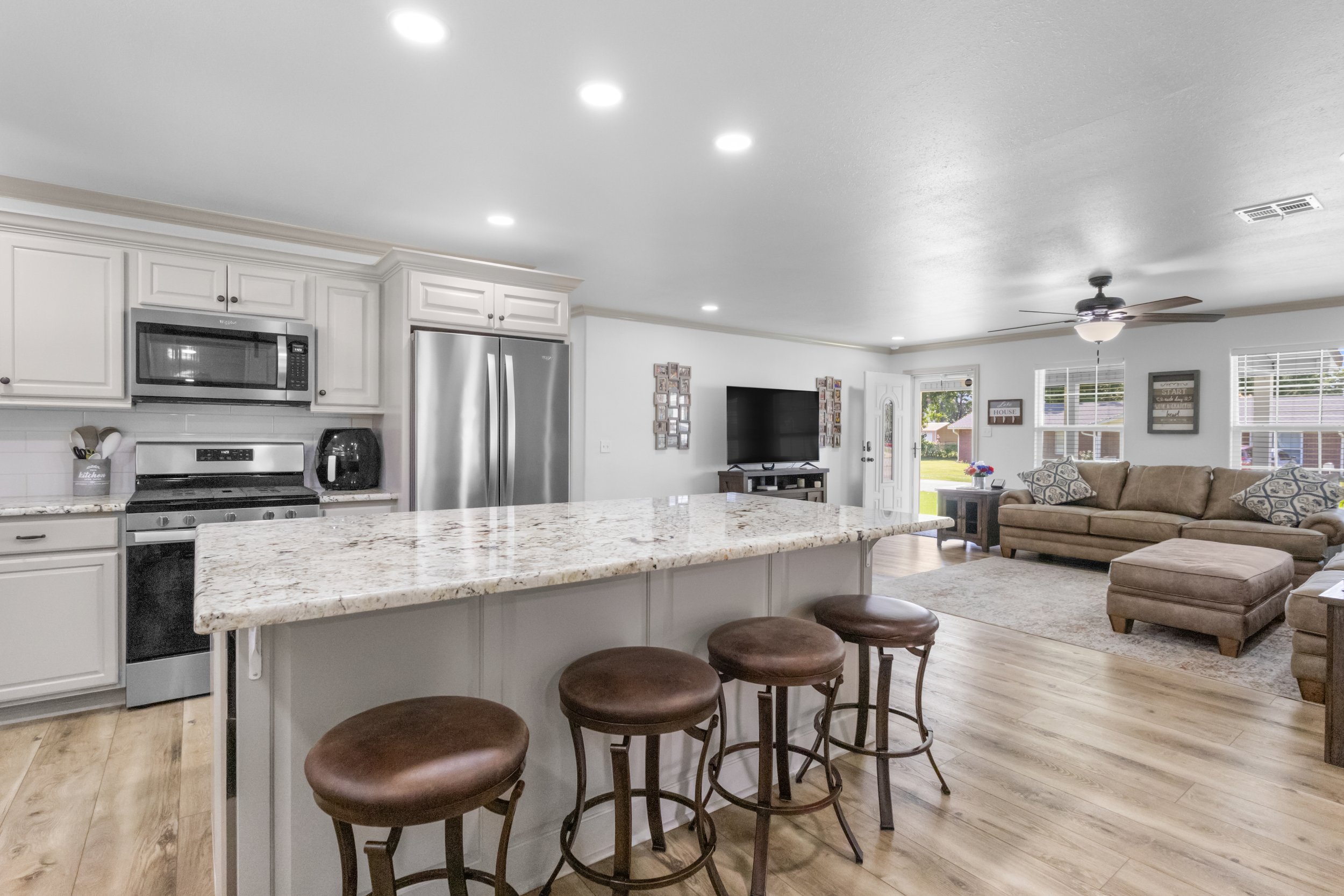Open-concept living room and kitchen with white cabinetry, stainless steel appliances, granite island, brown bar stools, beige sofa, wall-mounted TV, ceiling fan, and large windows with blinds.