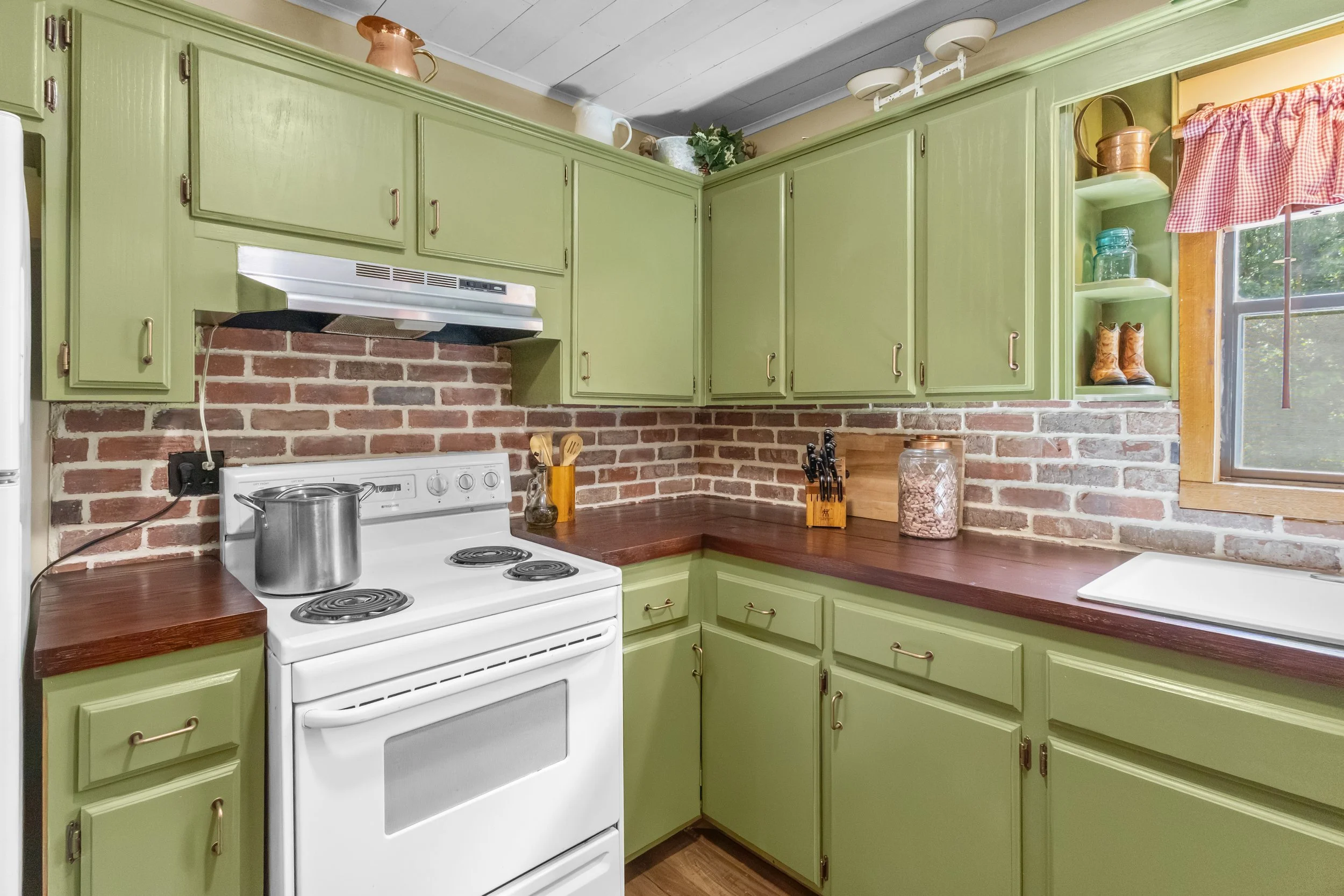 A kitchen with lime green cabinets, brick backsplash, and a white stove with a pot on top. There are wooden countertops, a window with red checkered curtains, and decorative jars and shoes on open shelves.
