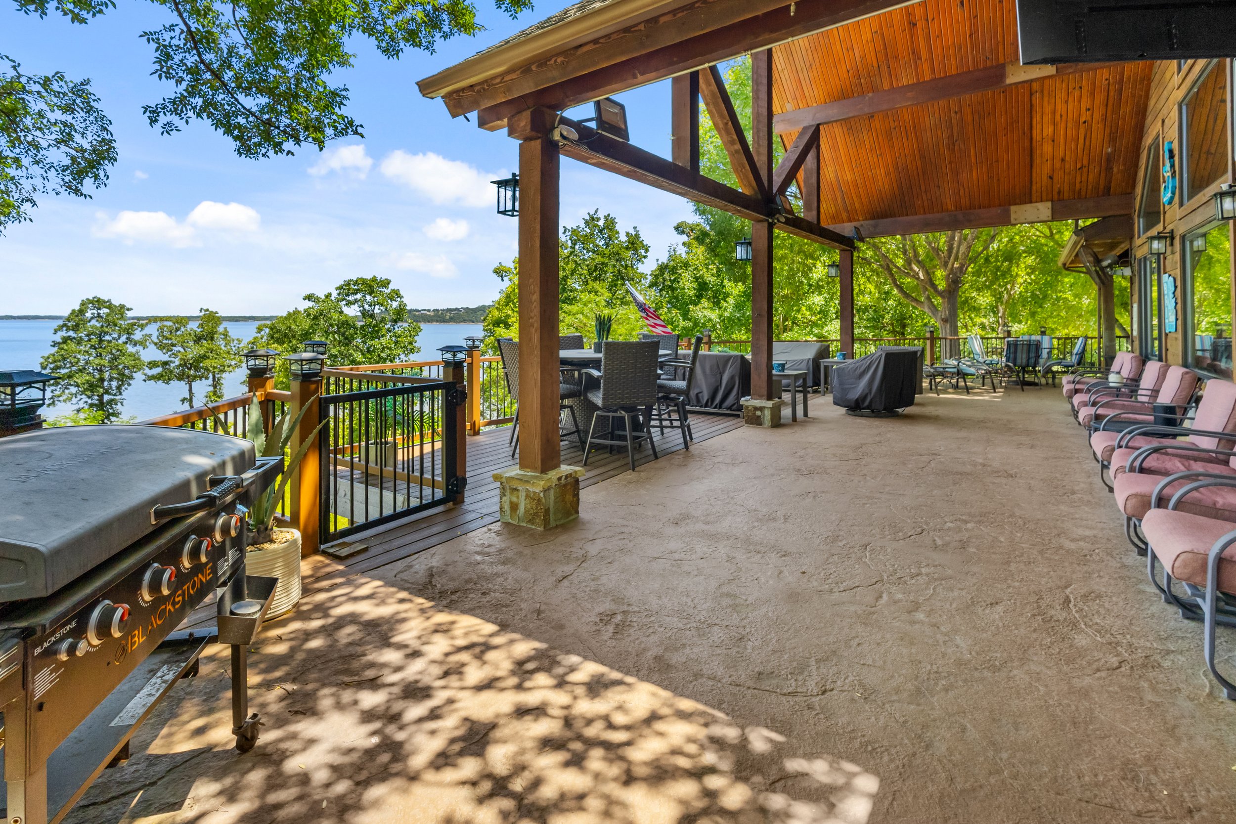 A spacious covered outdoor patio with a view of a lake, furnished with chairs, tables, and a BBQ grill, surrounded by green trees and a railing along the edge.