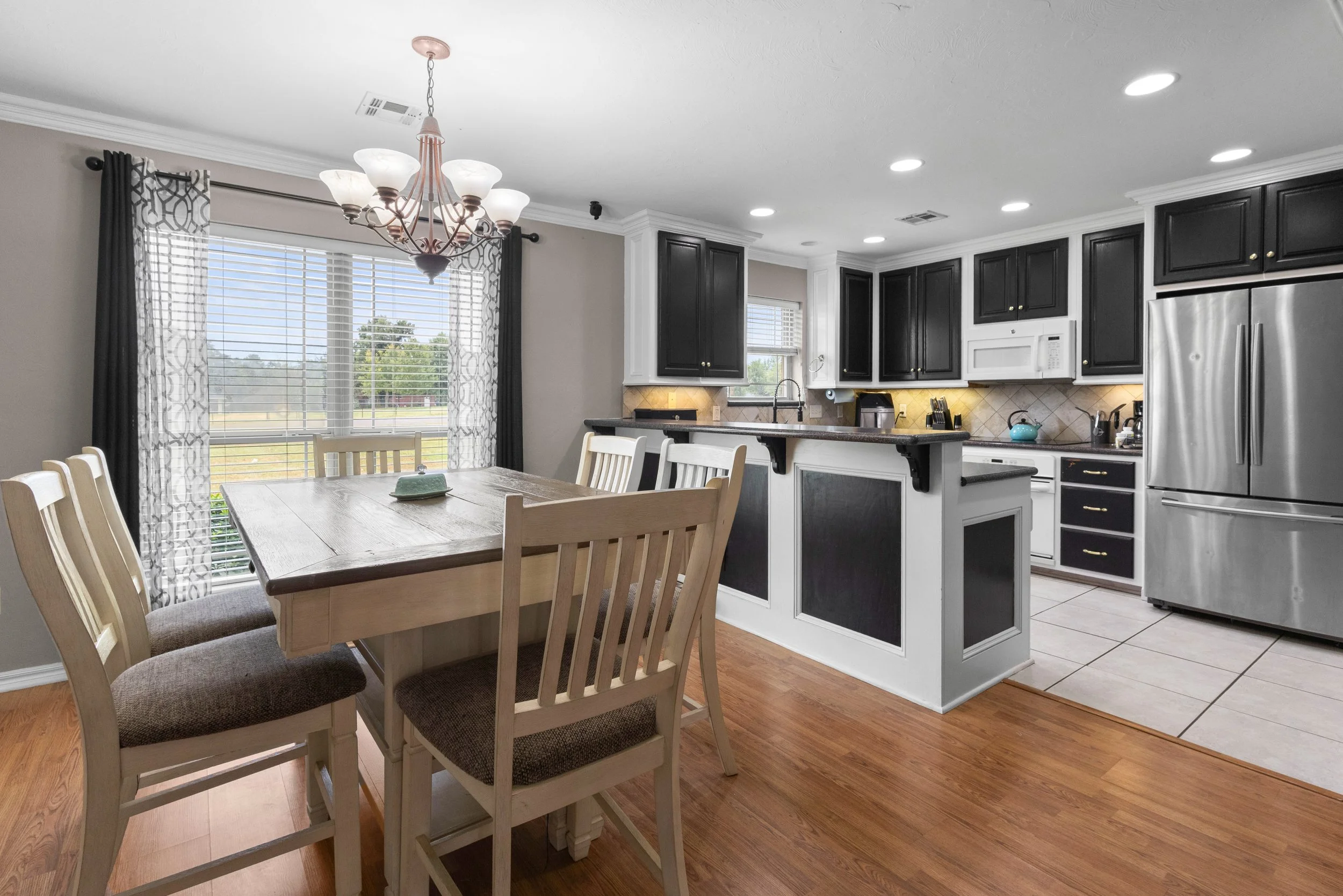 Dining area with a wooden table and six chairs in front of a window with patterned curtains. The kitchen has black cabinets, stainless steel refrigerator, microwave, and a black countertop island with a sink. Recessed lighting and a chandelier are also visible.