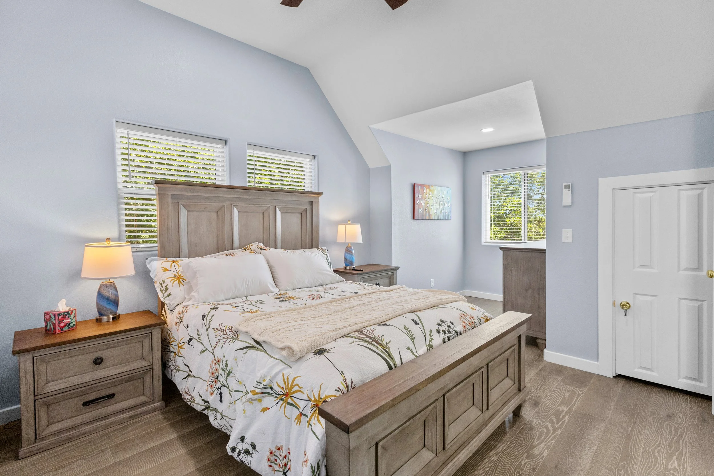 A cozy bedroom with a wooden bed frame, floral bedding, two nightstands with lamps, and three windows with white blinds, painted in light blue.