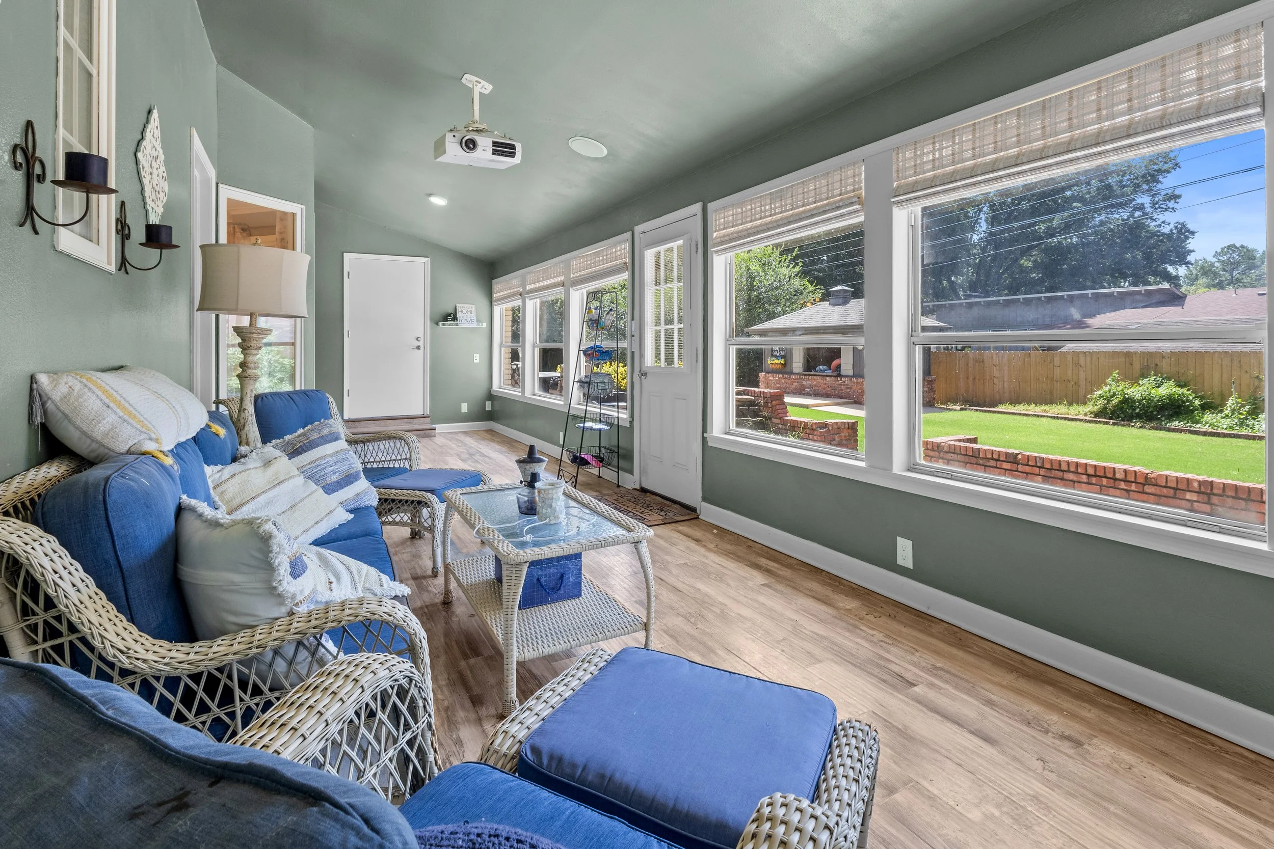 Sunroom with wicker furniture, blue cushions, and large windows offering a view of a backyard with a brick fence and lush greenery.