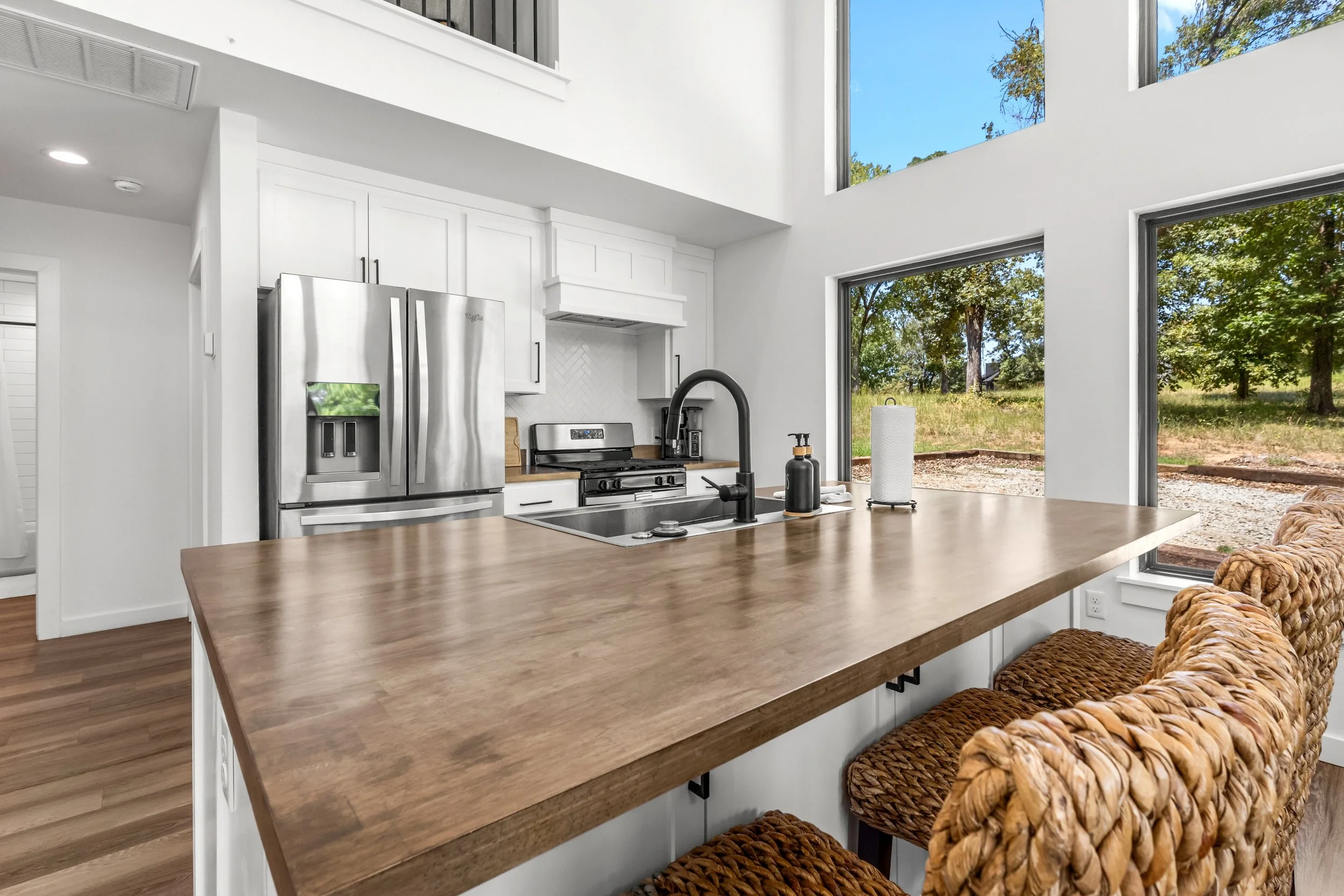 A modern kitchen with large windows showing a garden view, white cabinets, stainless steel refrigerator, and a wooden kitchen island with woven chairs.