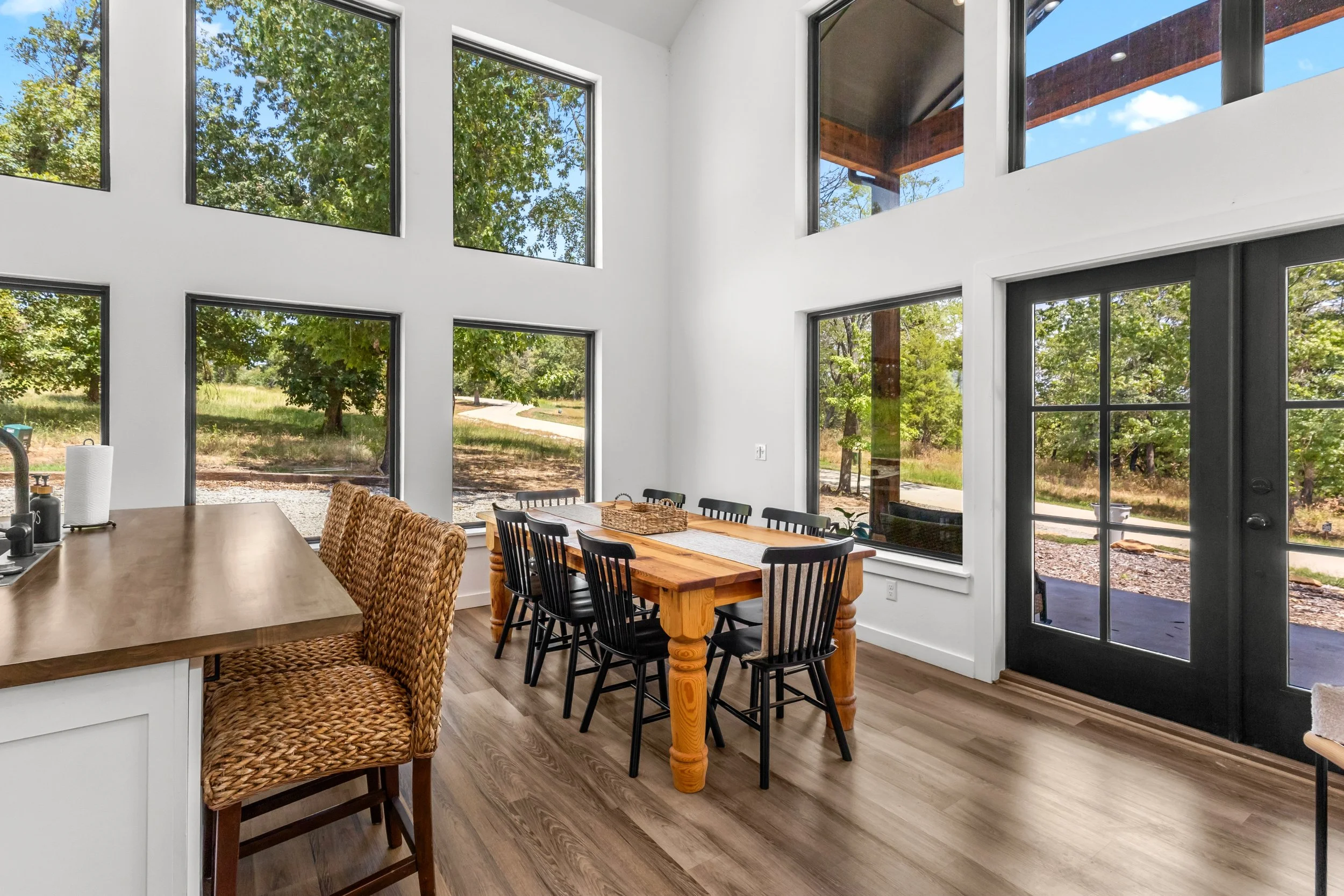 Interior view of a dining area with large windows showing a green outdoor landscape, a wooden dining table with black chairs, a woven chair, and a sliding glass door leading outside.