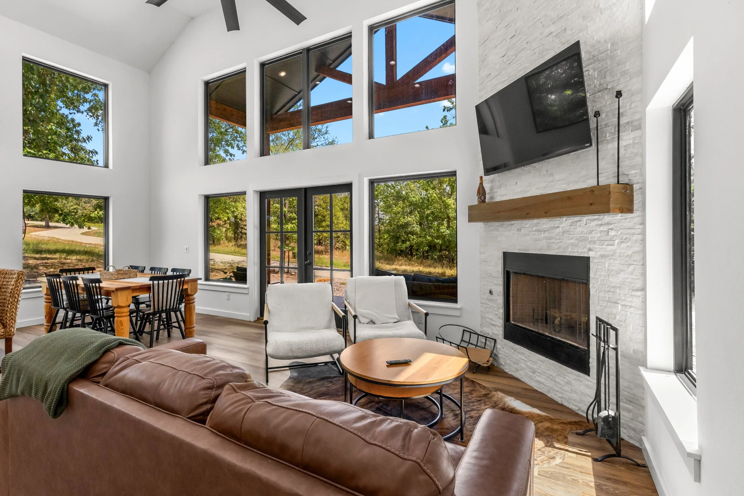 Interior of a modern living room with large windows, a white brick fireplace with a mounted flat-screen TV, a wooden mantel, and a seating area with a brown leather sofa, white armchairs, and a wooden coffee table.