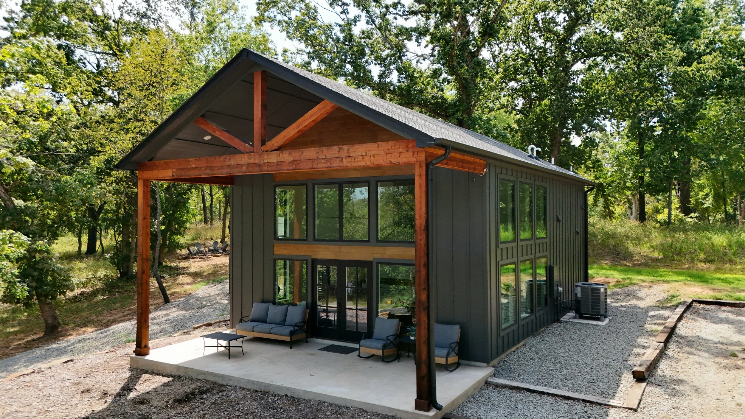 A modern two-story house with large windows, a covered porch, and outdoor seating, situated in a wooded area.