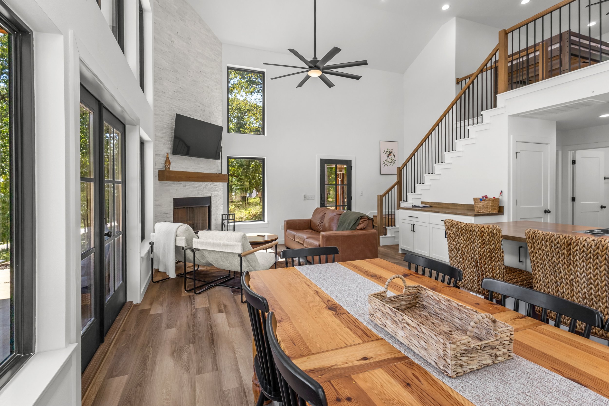 Open-concept living and dining area with large windows, white walls, vaulted ceiling, black ceiling fan, staircase with wooden handrail, fireplace with a wooden mantel, and a mix of modern and rustic furniture.