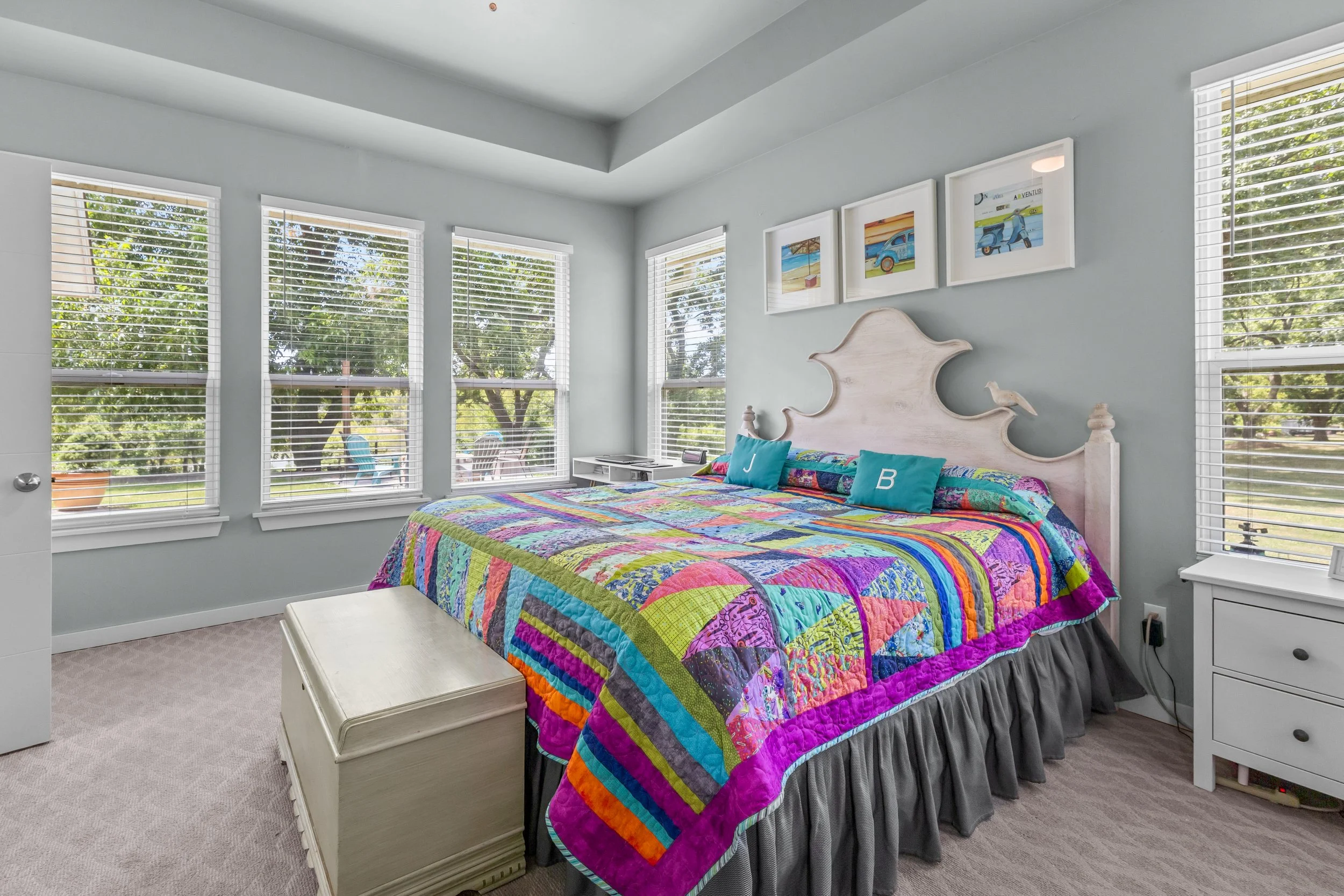 Bright bedroom with a colorful patchwork quilt on the bed, which has a white headboard. Large windows with white blinds offer a view of green trees outside. There are framed beach-themed pictures above the bed and a nightstand with a white finish on each side.
