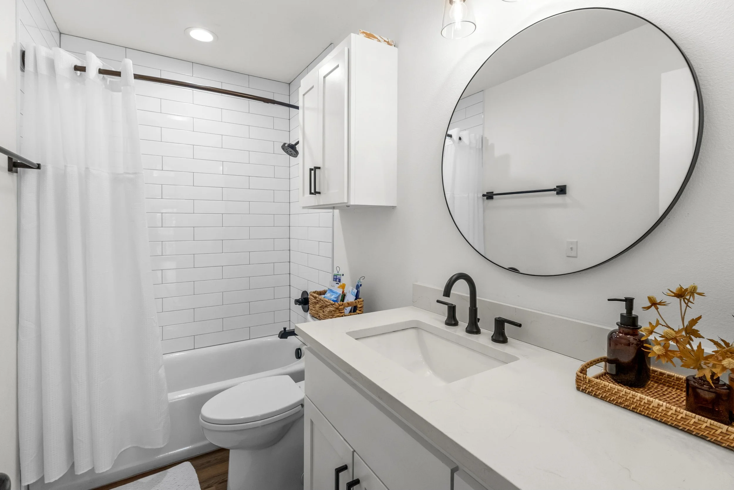 A modern bathroom with a bathtub and a shower, white subway tile walls, a round mirror, black fixtures, a white vanity with toiletries, a woven basket, and decorative plants.
