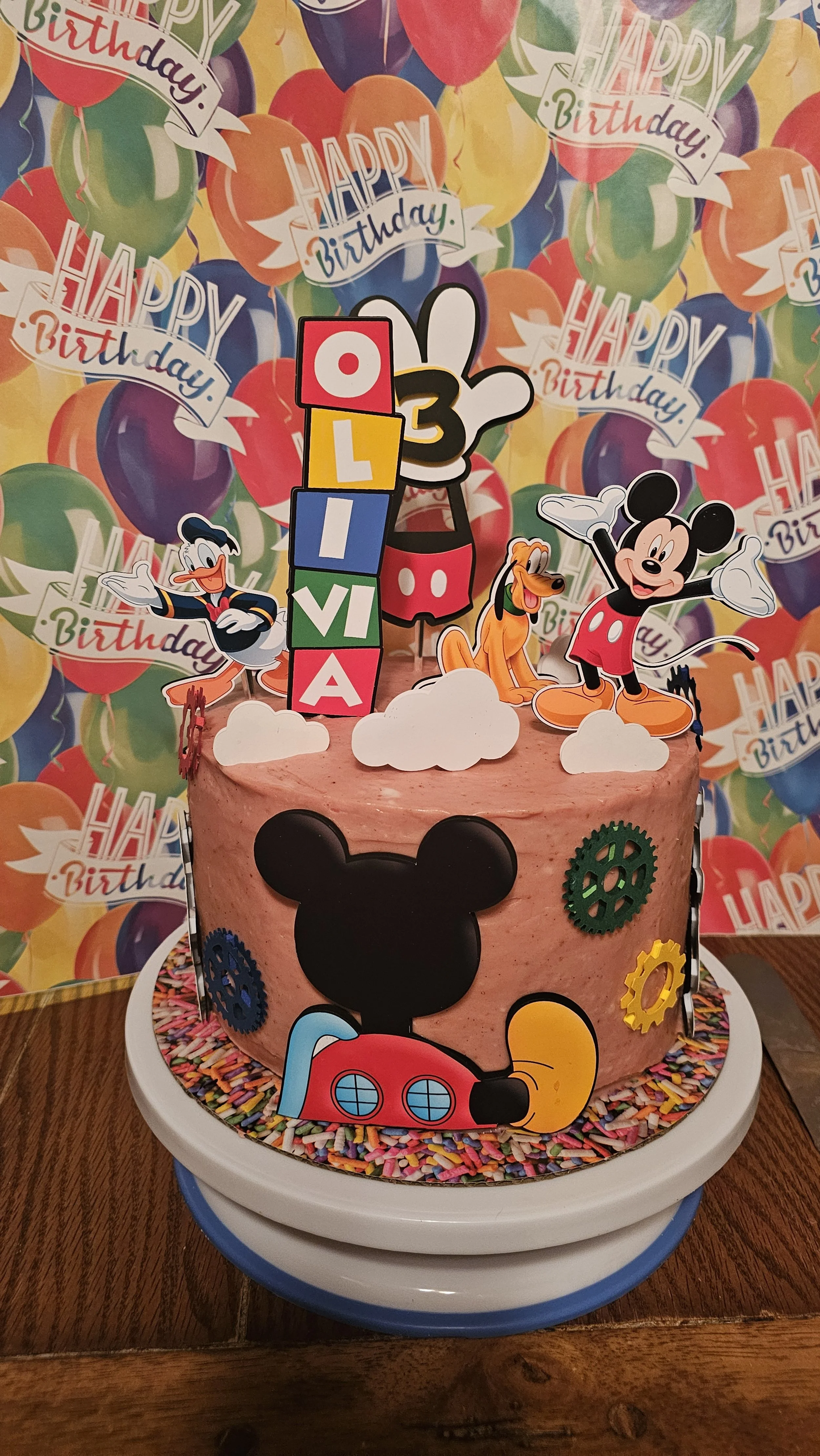 A birthday cake decorated with Mickey Mouse theme, featuring cake toppers of Mickey and friends, colorful balloons, and the name Olivia with the number 3 for a birthday celebration.