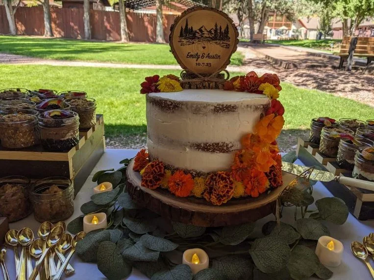 A semi-naked wedding cake decorated with orange, yellow, and red flowers, featuring a wooden topper with trees and the names Emily and Austin with the date 10.7.23, placed on a table with greenery and small candles, outdoors in a park setting.