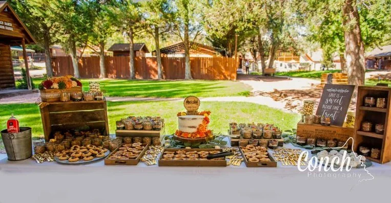 Dessert table at an outdoor wedding event with cookies, cupcakes, and a cake, decorated with autumn-themed items, on a white tablecloth in a backyard setting