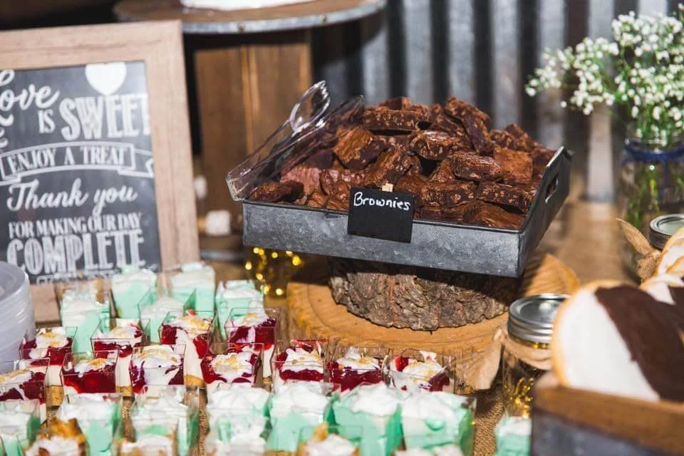 Tray of brownies labeled 'Brownies' on a rustic wood stand, with individual dessert cups with whipped cream and red sauce, and jars with snacks, on a table with a floral arrangement in the background.