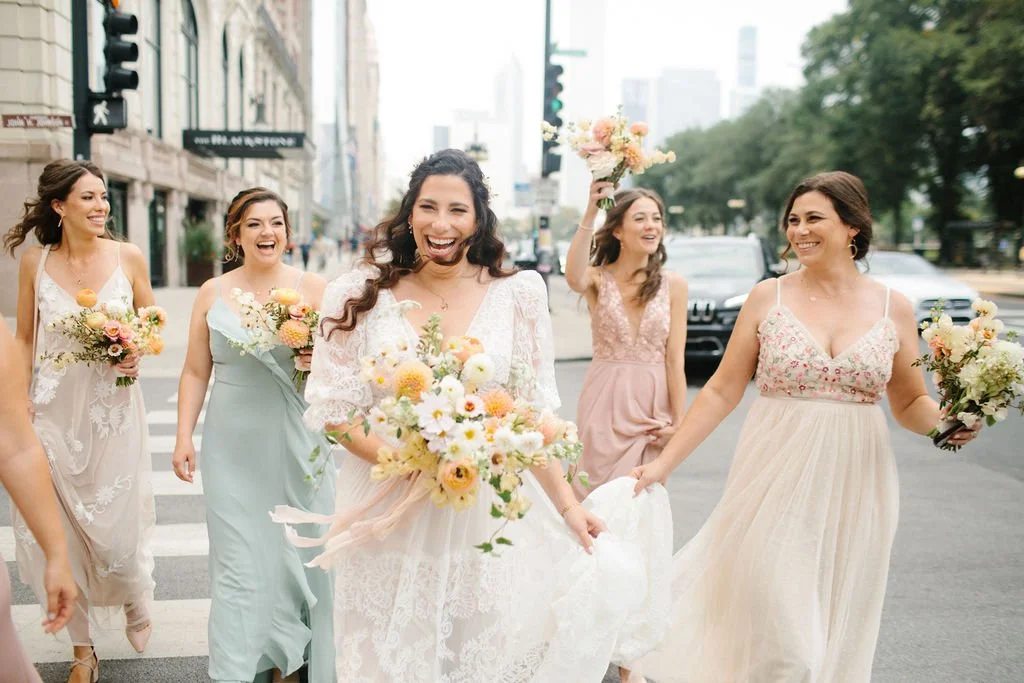 Mehrere Frauen in eleganten Kleidern, die ein Brautpaar bei einer Hochzeit auf der Straße begleiten, alle lachen und halten Blumen.