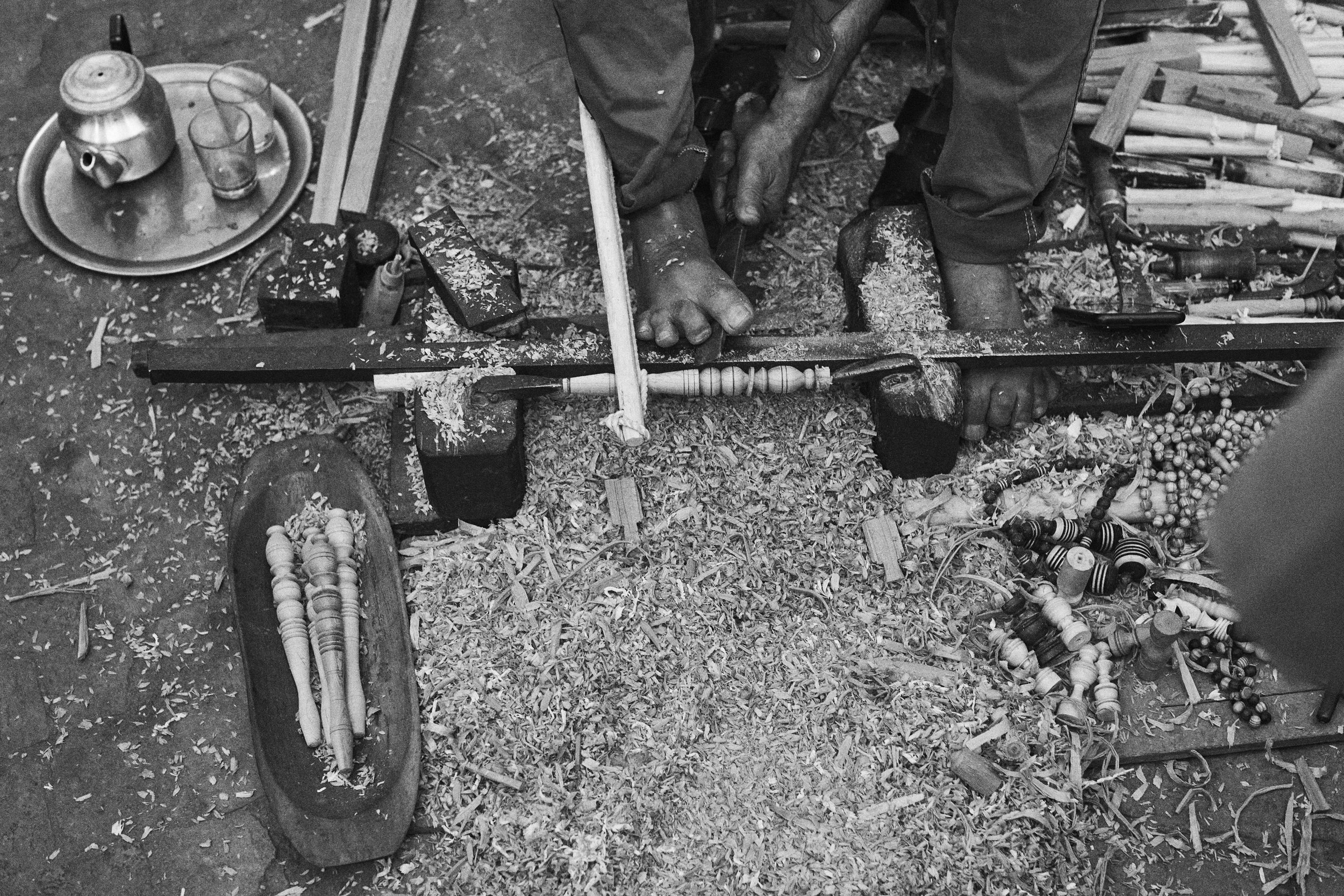 Black and white photo of a craftsman in the Medina of Marrakech, Morocco, using his feet and hands to carve wood on a traditional lathe, surrounded by wood shavings, tools, and finished spindles.