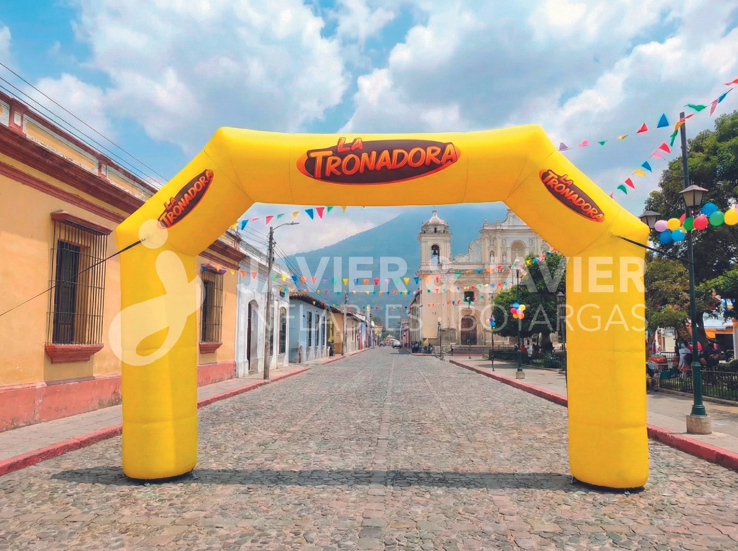Arco inflable amarillo con la marca La Tronadora en una calle de un pueblo, decorada con banderines de colores y globos.