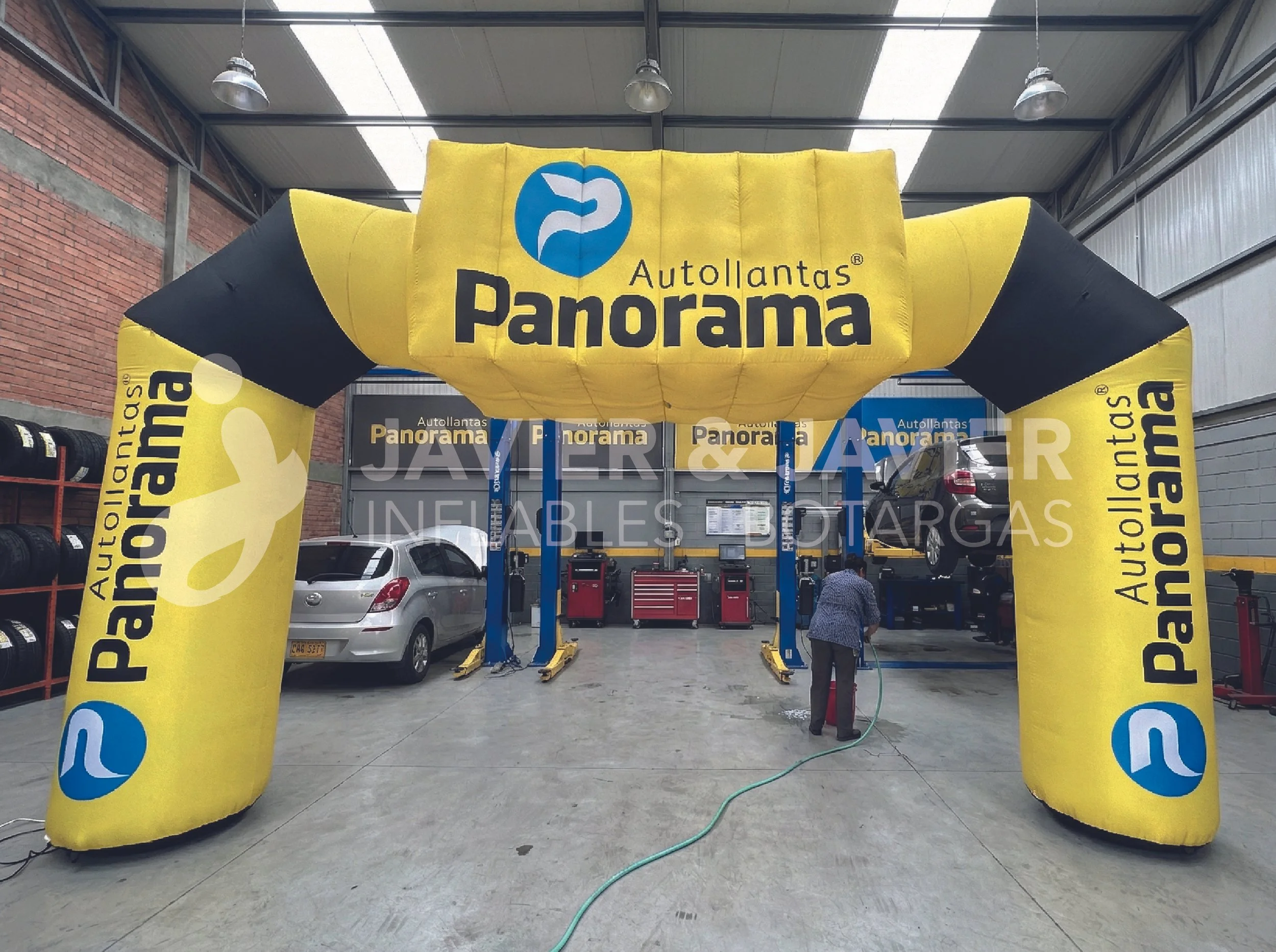 Un taller de autos con un arco inflable amarillo y negro que dice 'Autollantas Panorama'. Hay coches elevados para servicio en un espacio cerrado con paredes de bloques de ladrillo y metal, y una persona lavando un auto con una manguera.