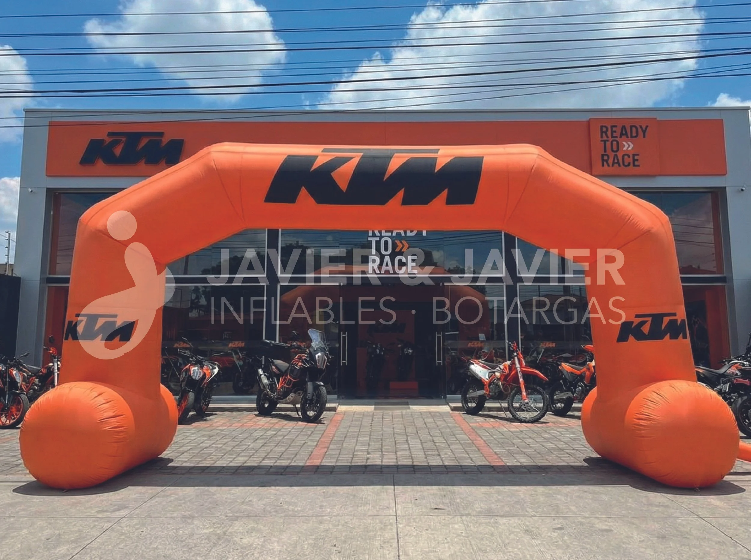 Entrada de tienda de motocicletas con un arco inflable naranja con logotipo KTM y cartel que dice 'Ready to Race' en el exterior, con varias motocicletas exhibidas frente a la tienda