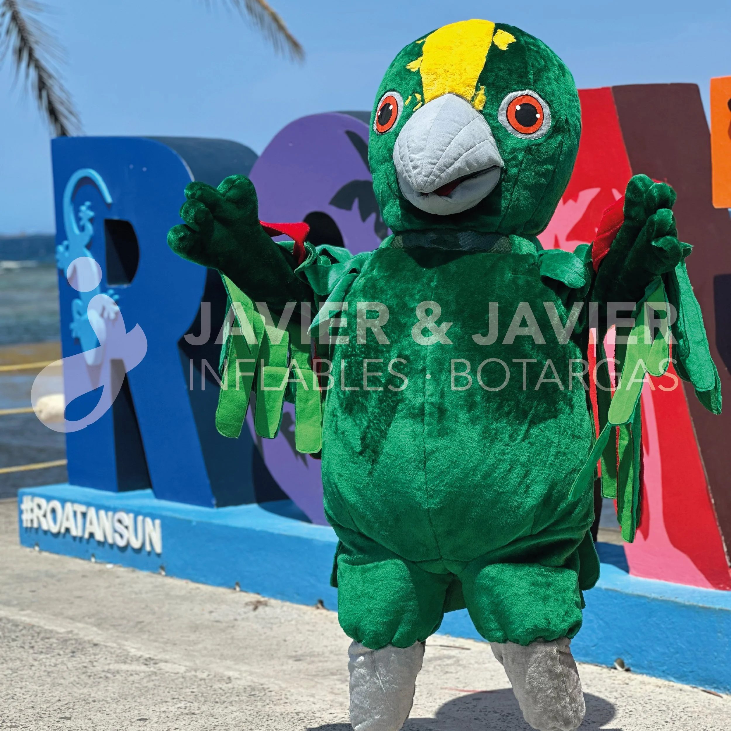 Mascota de carnaval en forma de loro verde con detalles en amarillo y gris, con fondo de letras grandes de colores y una playa al fondo.