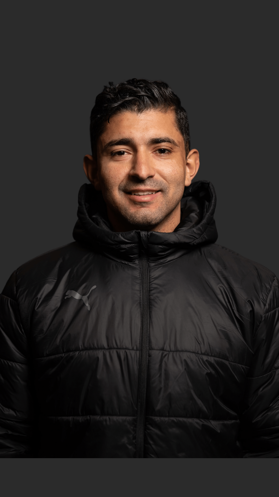 Young man smiling wearing a black puffer jacket with a Puma logo, against a dark background.