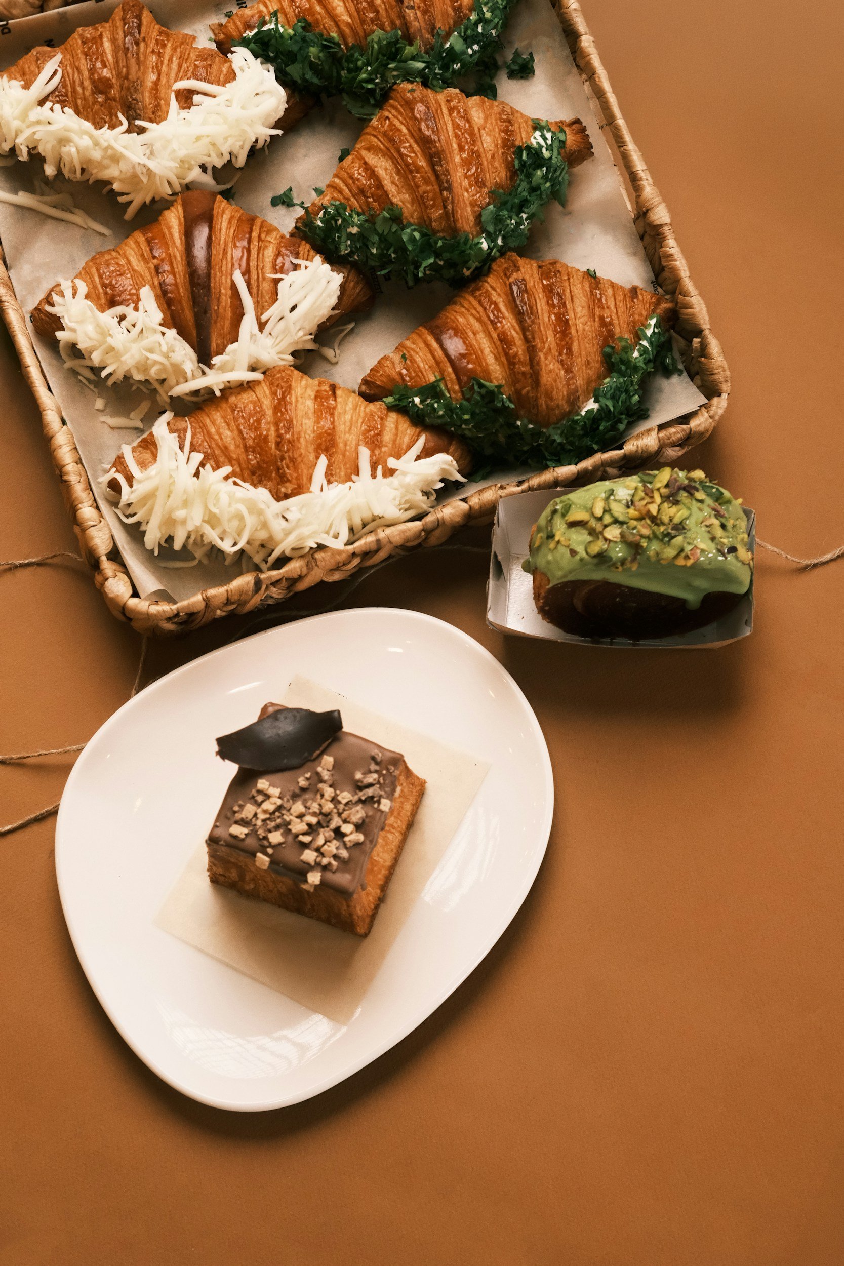 A basket containing croissants topped with chopped parsley and shredded cheese. Next to the basket, there is a piece of chocolate-covered cake with chopped nuts on top placed on a white plate.