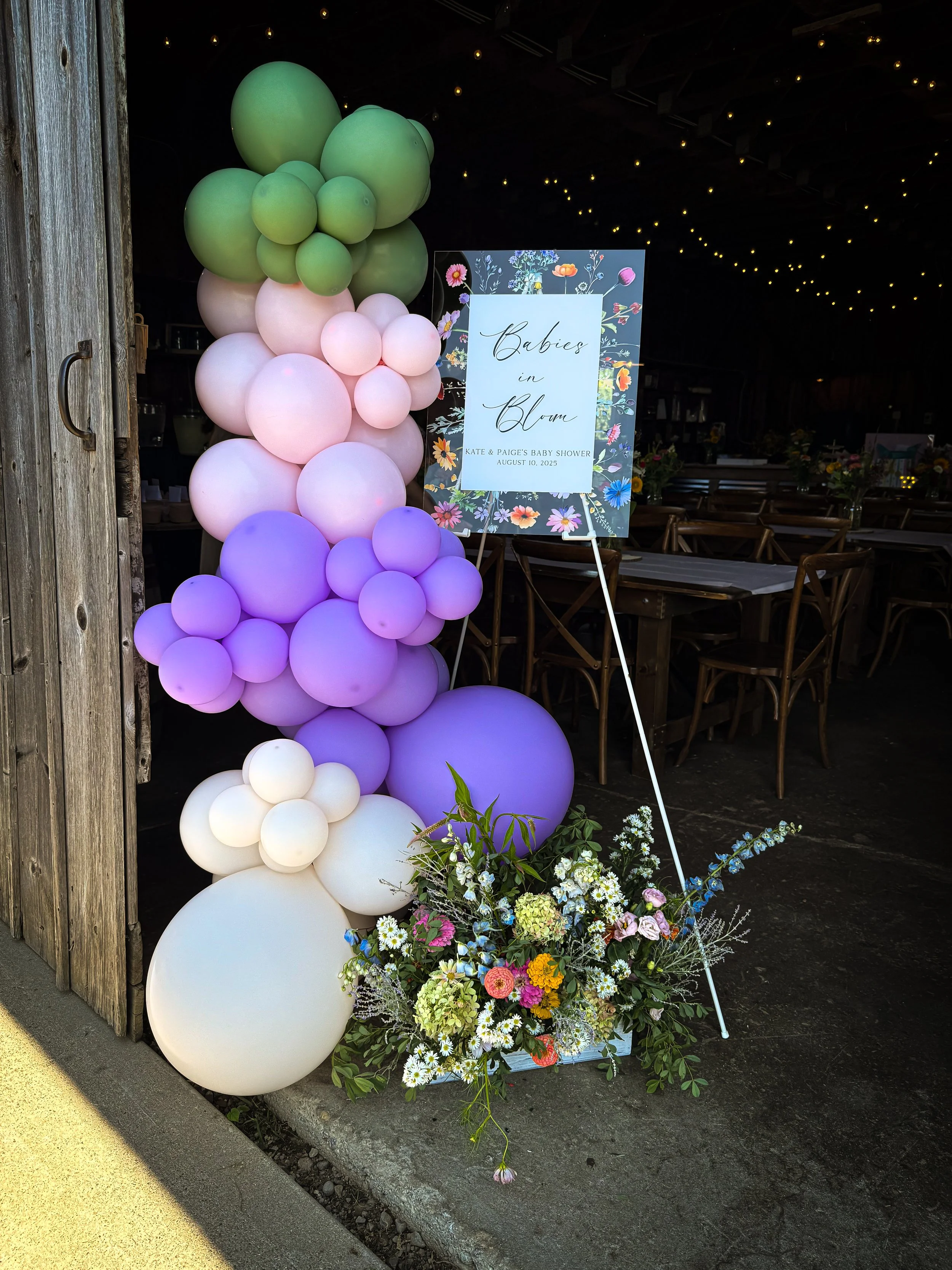 Decorative balloon arrangement with pastel-colored balloons in green, pink, purple, and white, alongside a floral sign and flower arrangement for a baby shower event.