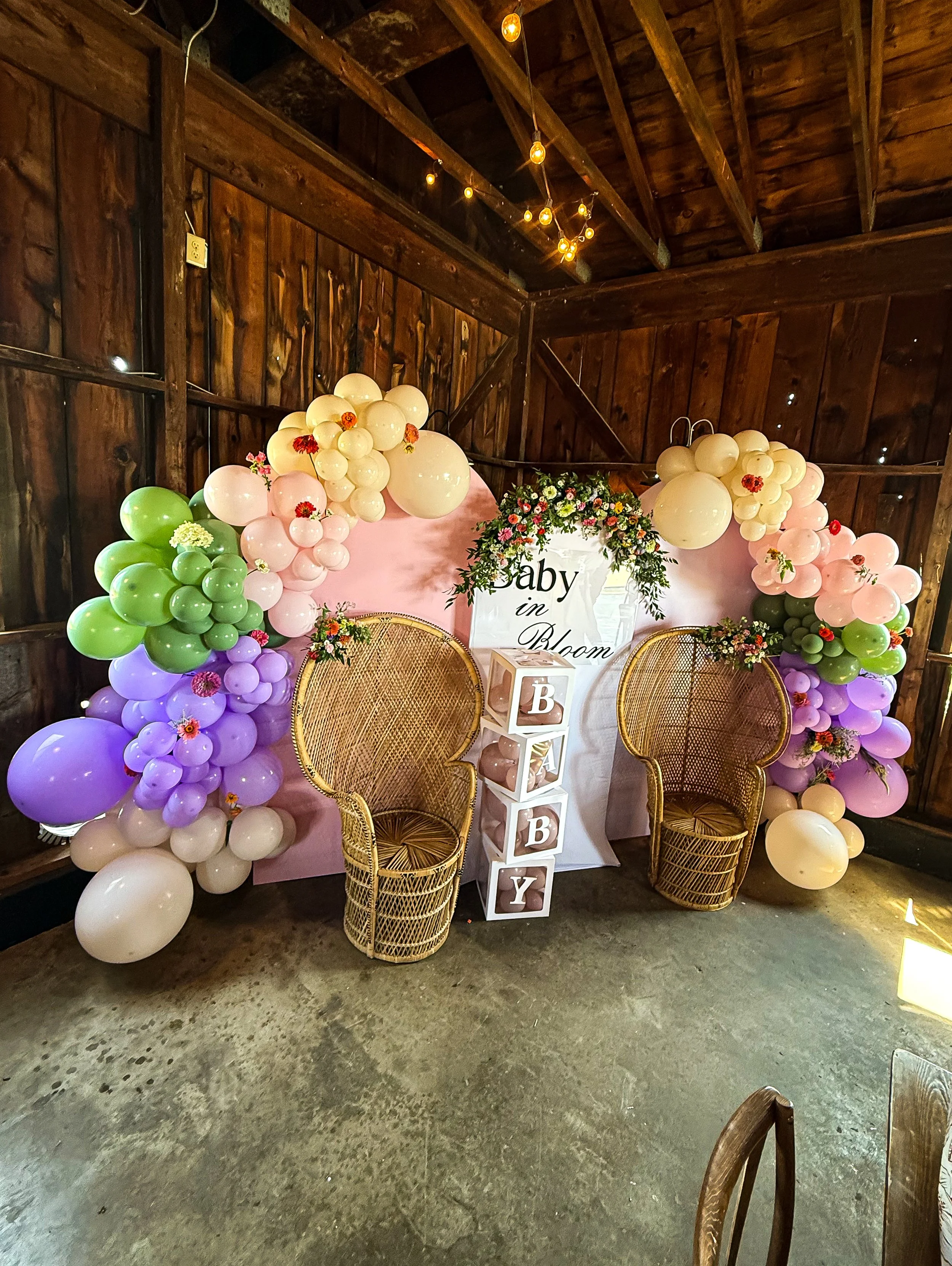Decorative baby shower setup with ballon arch, two wicker chairs, and a purple and green color theme.