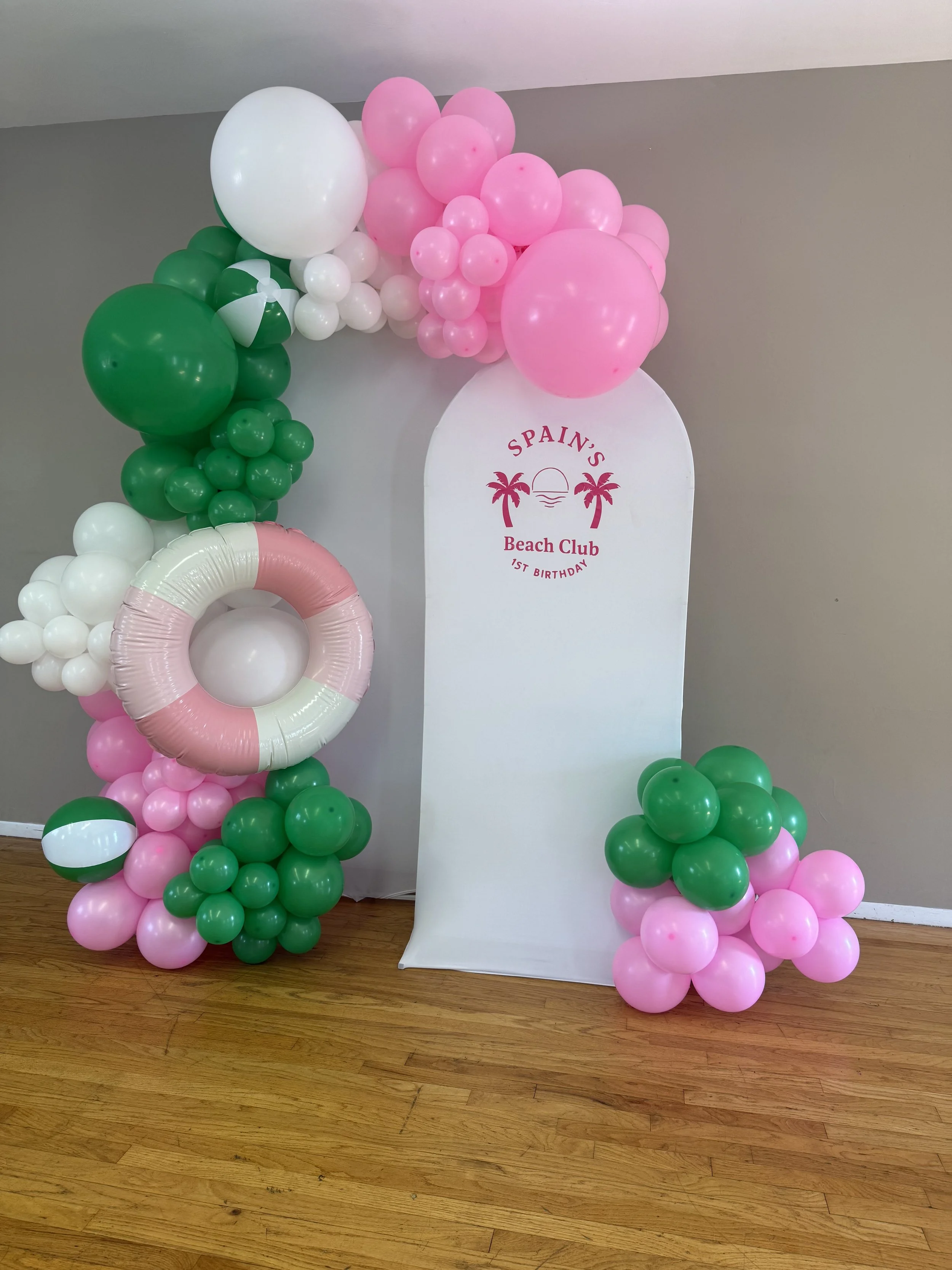 Decorative balloon arrangement in pink, green, and white colors with a pink and white lifebuoy and a beach club sign for Spain's Beach Club 1st birthday party.