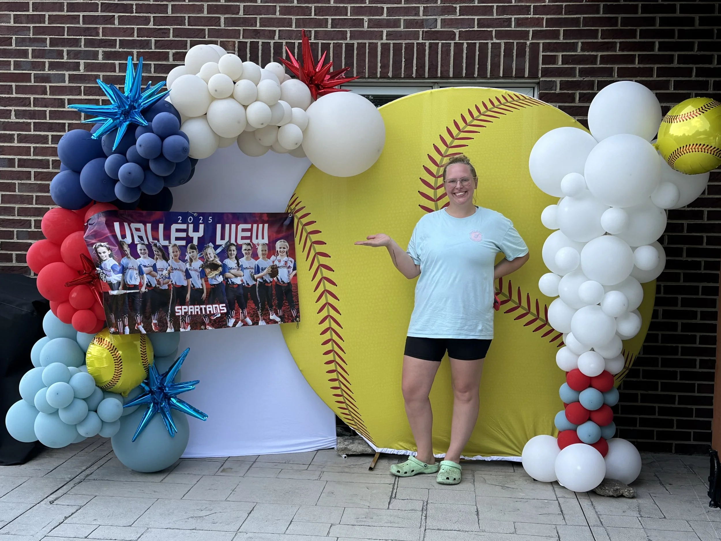 Softball-themed balloon backdrop with red, white, and blue balloon garland, metallic star balloons, and a large yellow softball display for the Valley View Spartans 2025 team celebration