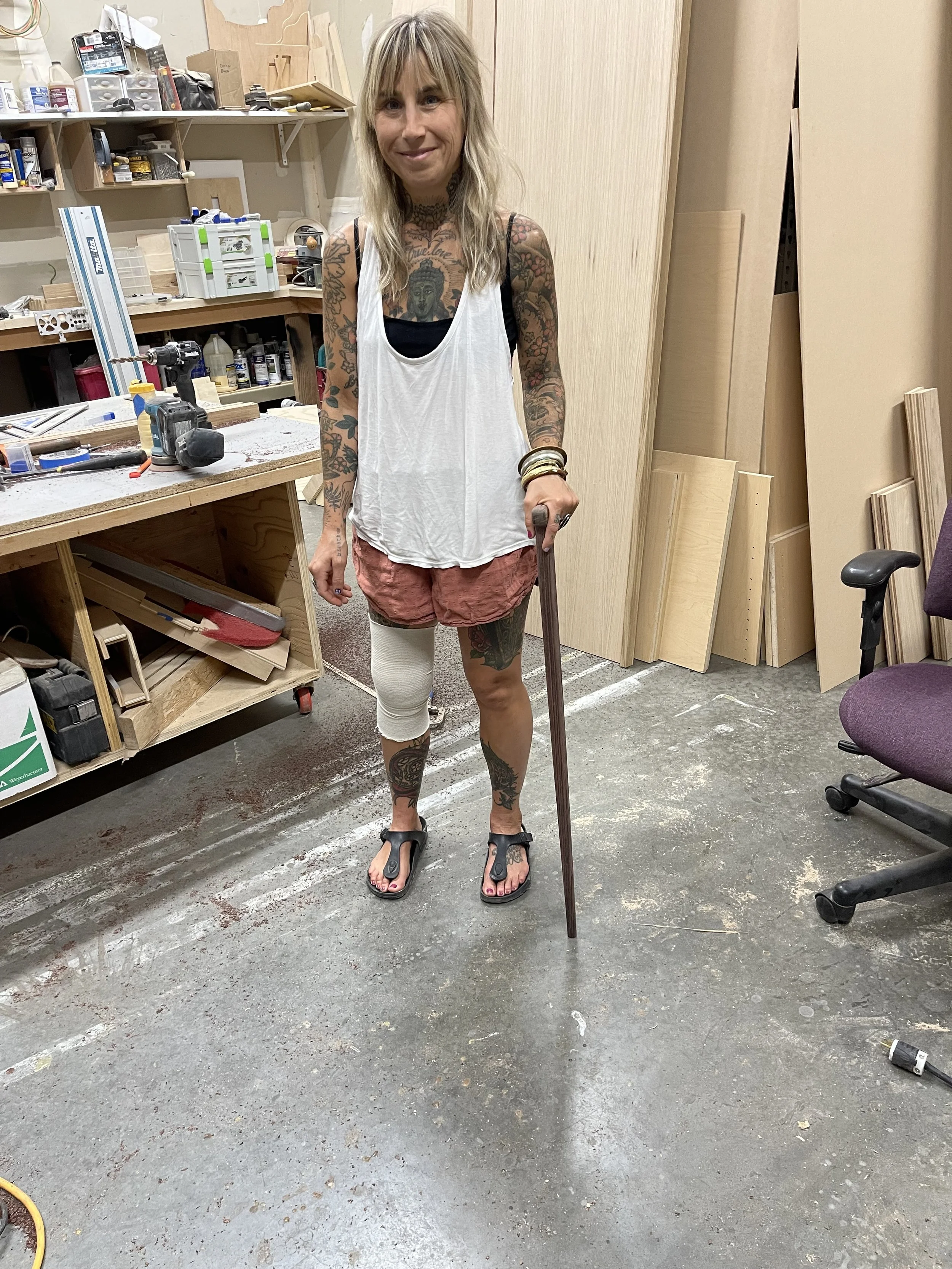 A woman with tattoos on her arms and legs, wearing a white tank top, pink shorts, and black sandals, stands in a woodworking workshop holding a cane or walking stick.