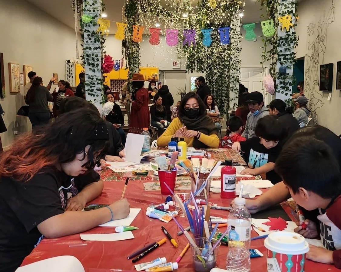 A group of children and adults participating in a holiday-themed arts and crafts event in a decorated indoor space with banners, string lights, and foliage.
