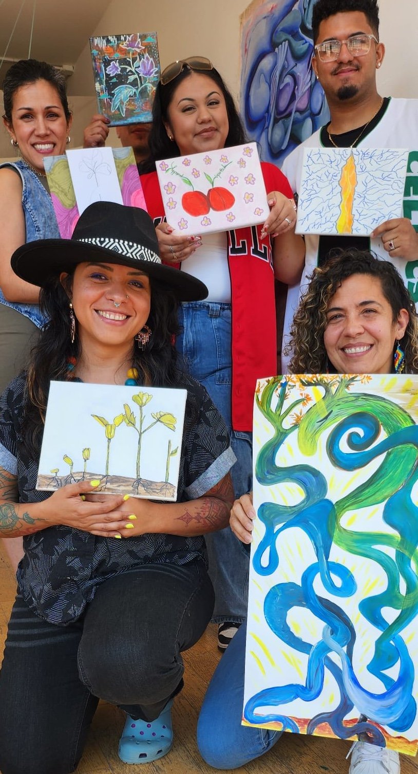 Group of six people smiling and holding colorful paintings, posing indoors against a wall with artwork.