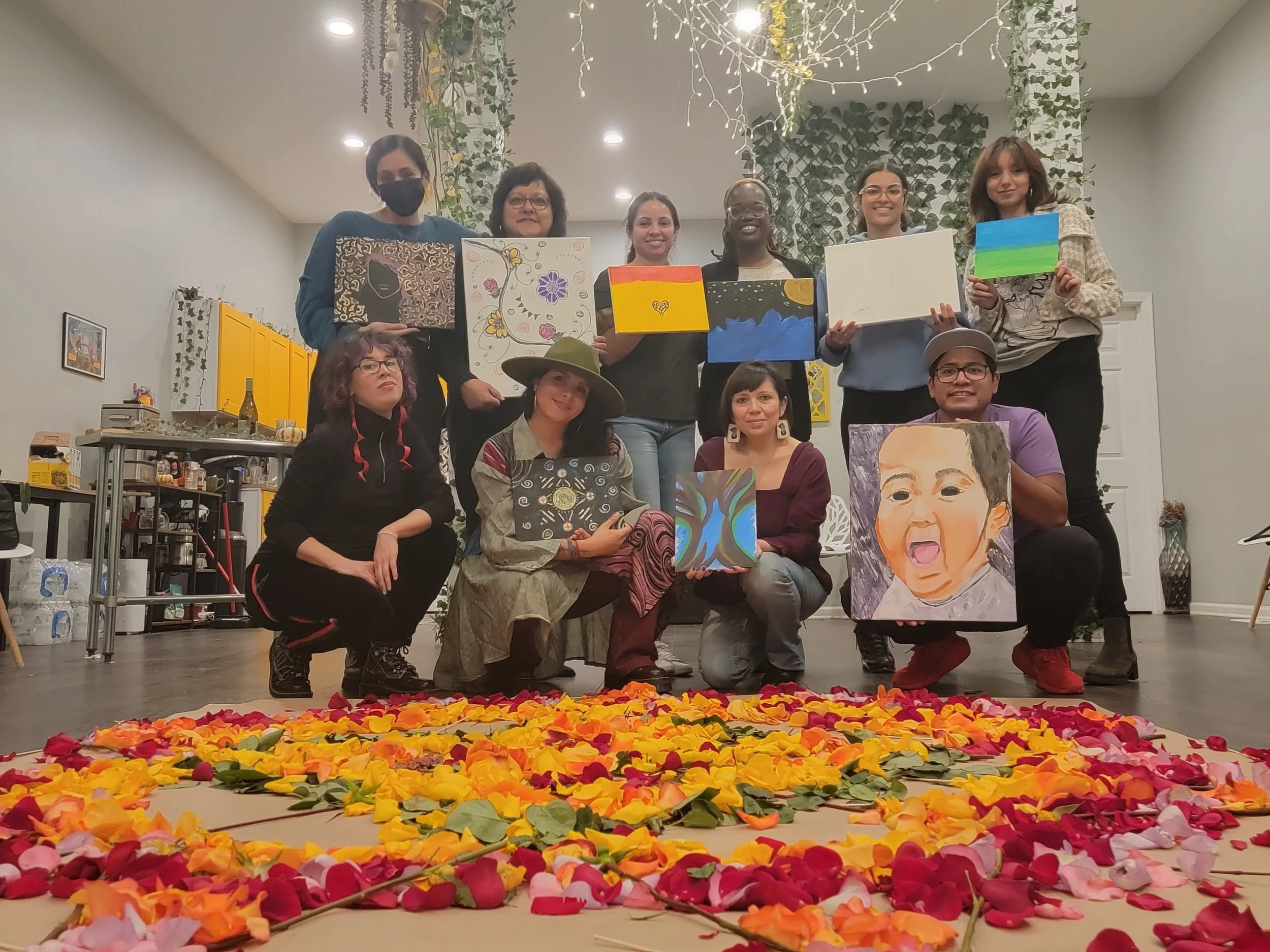 Group of people posing with paintings they created, surrounded by colorful flower petals on the floor, indoors with hanging plants and lights.