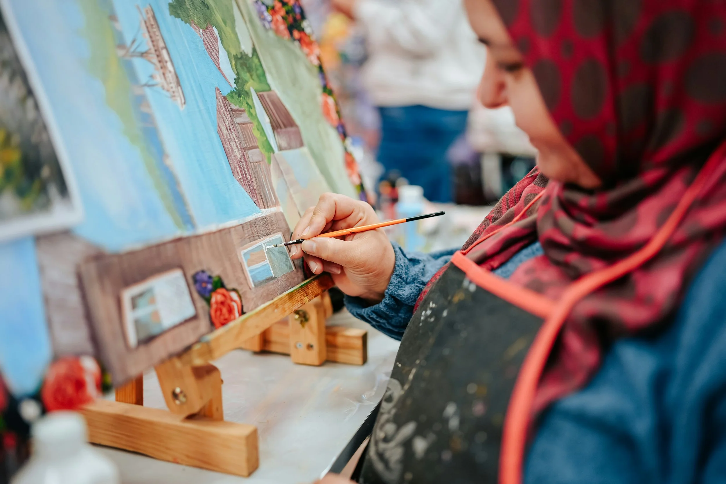 Woman painting a colorful landscape on a canvas, wearing a headscarf and apron, with art supplies nearby.