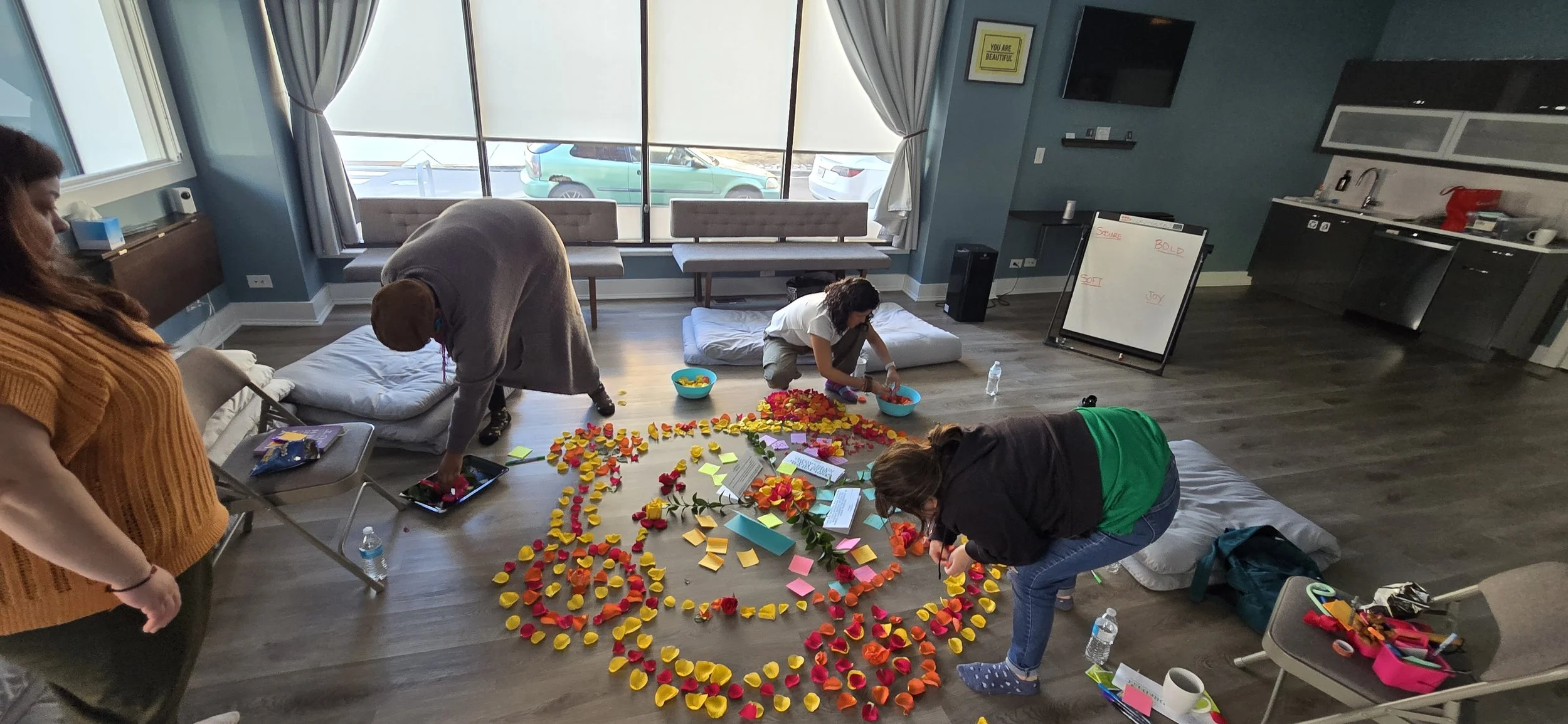 People arranging yellow, red, orange, and pink flower petals and sticky notes on the floor to create a pattern or message in a room with large windows, a television, and a kitchenette.