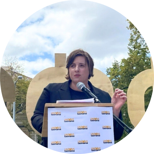 Portrait of Nicole McPherson, a woman with shoulder-length brown hair speaking at a lectern with her hand raised.