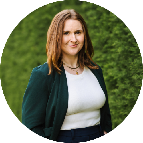 A light-skinned person with shoulder length reddish brown hair and wearing a dark green blazer and white top. They are standing in front of a green hedge and looking directly at the camera with a slight smile.