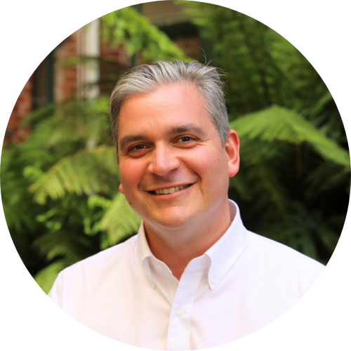 Portrait of a man's face and shoulders, wearing a white business shirt in front a leafy background.