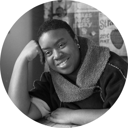 Maxine Beneba Clarke, a Black woman with a short afro and earrings , is smiling, sitting at a table resting her head on her hand.