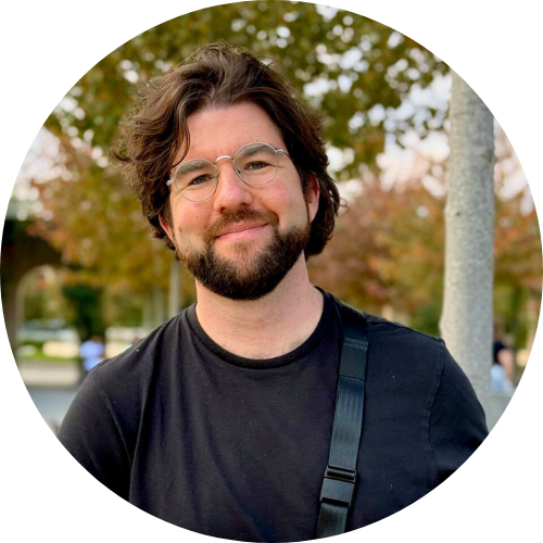 Viktor: a man with medium-length wavy brown hair, a short beard and round glasses standing outdoors, smiling softly at the camera.