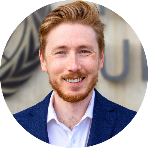 Jason is smiling at the camera, he is in a blue blazer and a buttoned-up white shirt in front of the UN