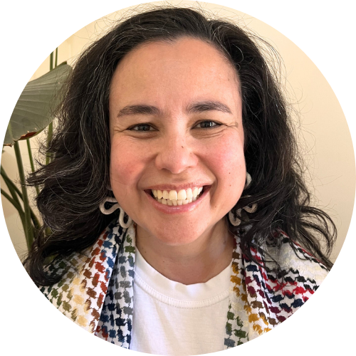 Sarah is a biracial woman with mostly black hair that has some grey running through it. She is smiling, looking directly at the camera. She is wearing white curly earrings and a colourful keffiyeh. There is a green plant behind her.
