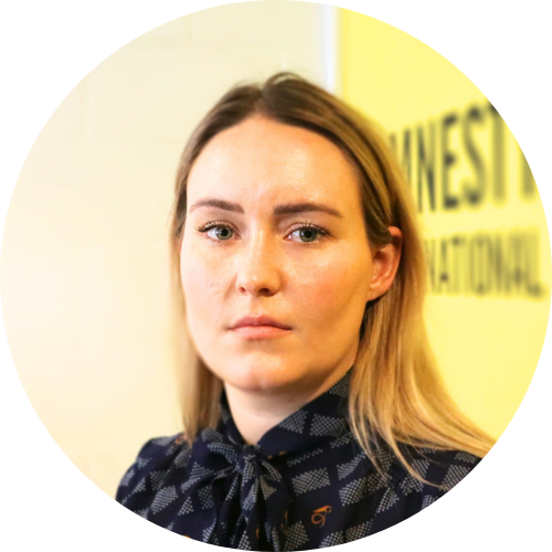 Portrait of Nikita White, facing the camera in front of a yellow Amnesty International wall