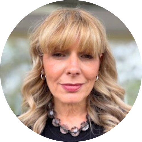 Headshot of Nina, a woman with shoulder-length warm blonde wavy hair and fringe, wearing a black top and grey beaded necklace, photographed outdoors against a soft green background.